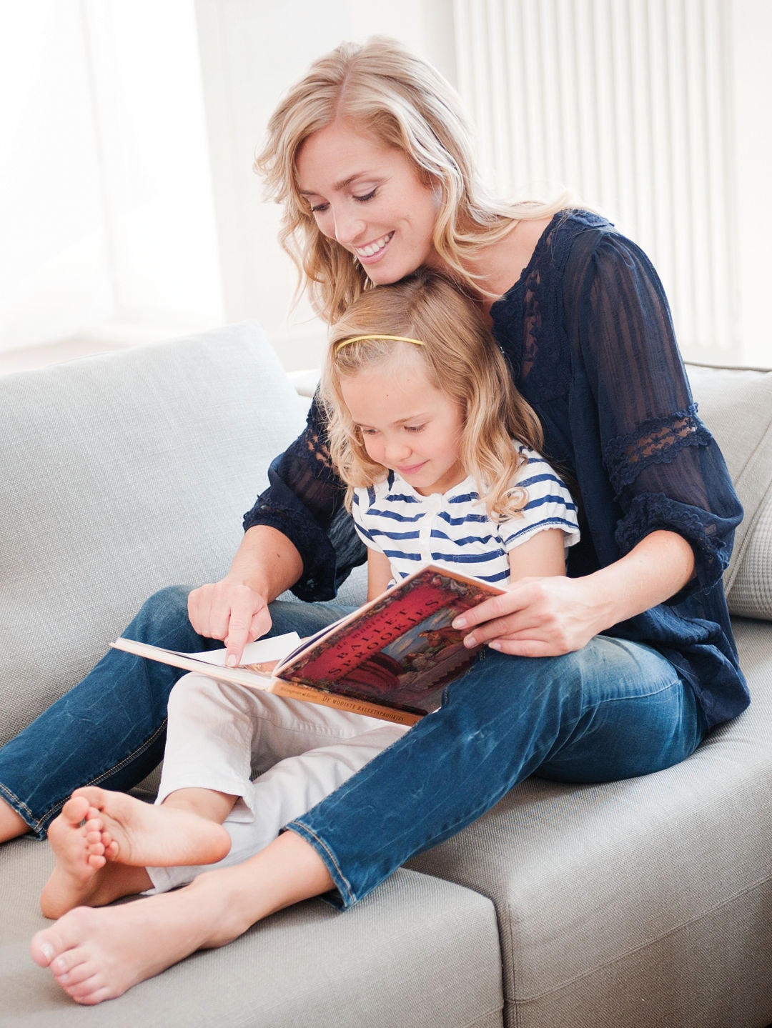 Mãe com uma filha pequena sentada no seu colo observam um livro no sofá